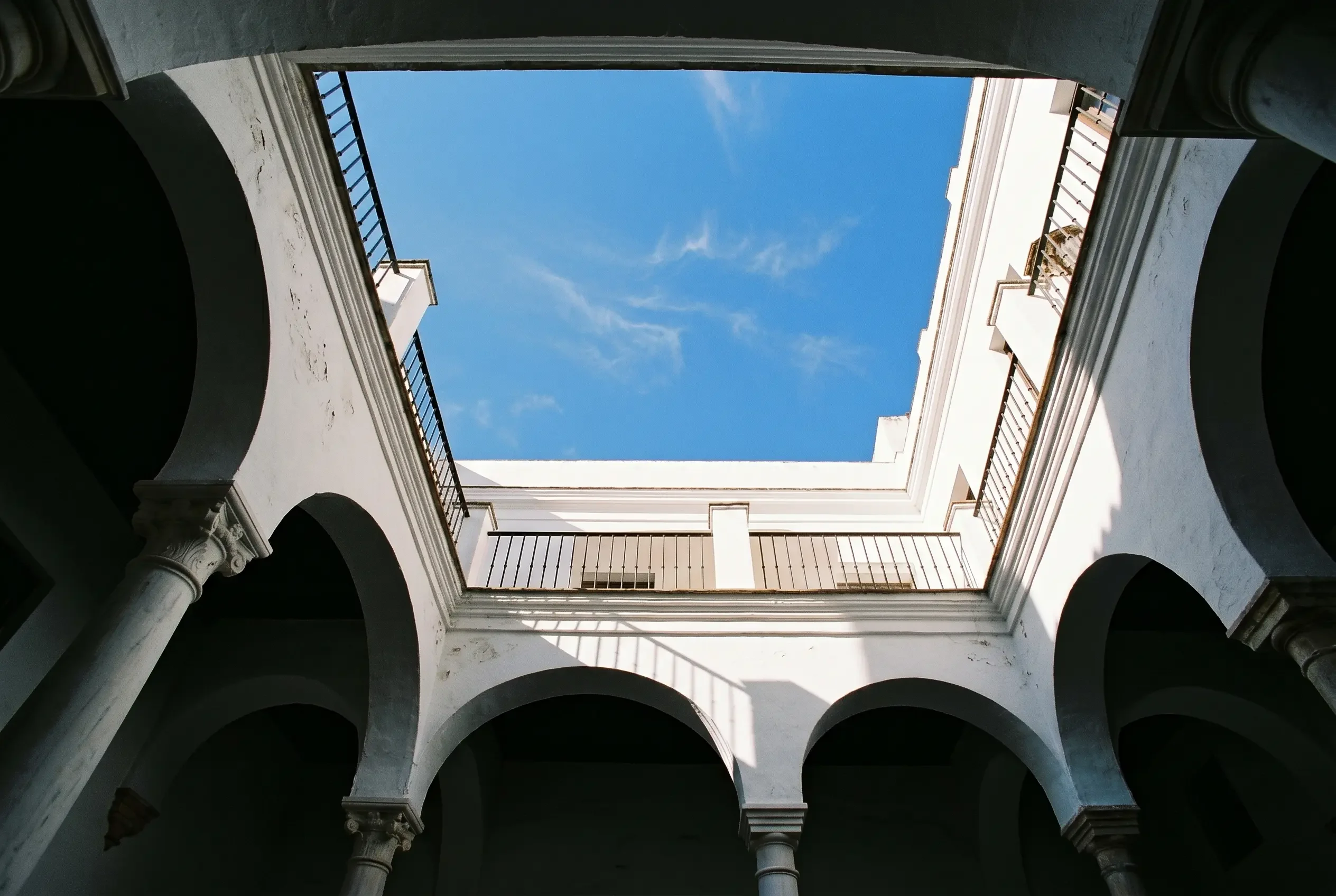 Vista cenital desde el interior de un patio andaluz hacia el cielo, con galerías en sombra y el rectángulo de cielo iluminado arriba.